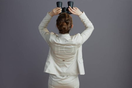 Foto de Visto desde detrás de una trabajadora vestida con un traje de negocios liviano con prismáticos mirando hacia arriba sobre fondo gris. - Imagen libre de derechos