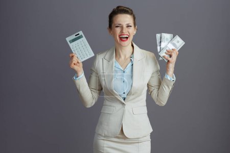 Téléchargez les photos : Heureux élégant femme d'âge moyen employé dans un costume d'affaires léger avec calculatrice et dollars paquets d'argent sur fond gris. - en image libre de droit