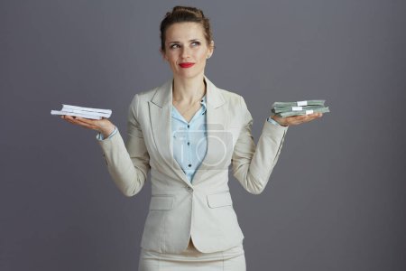 Foto de Empleada femenina de moda pensativa en un traje de negocios ligero con paquetes de dinero en dólares y paquetes de dinero en euros evaluando aislado en gris. - Imagen libre de derechos
