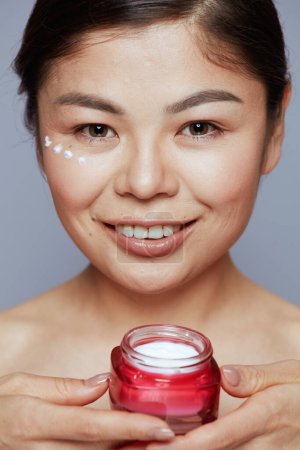 Foto de Retrato de mujer asiática joven con frasco de crema facial y crema facial en la cara sobre fondo azul. - Imagen libre de derechos