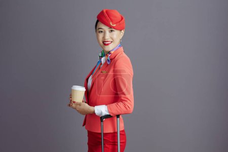 Foto de Sonriente moderno azafata asiática mujer en falda roja, chaqueta y sombrero uniforme con taza de café y bolsa de ruedas sobre fondo gris. - Imagen libre de derechos