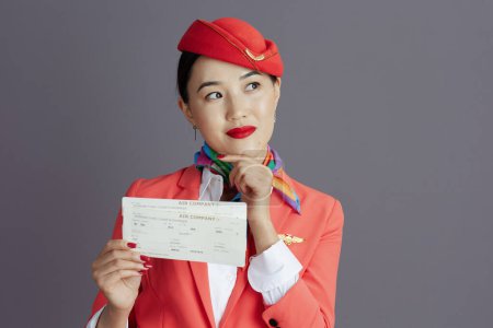 Photo for Pensive elegant flight attendant asian woman in red skirt, jacket and hat uniform with flight tickets looking at copy space isolated on grey. - Royalty Free Image