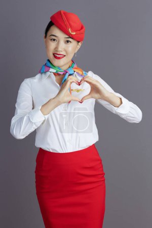 Photo for Smiling modern asian female flight attendant in red skirt and hat uniform showing heart shaped hands against gray background. - Royalty Free Image