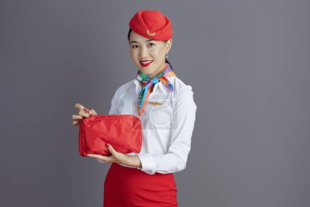 Photo for Happy stylish flight attendant asian woman in red skirt and hat uniform with first aid kit isolated on gray background. - Royalty Free Image
