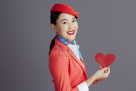Photo for Smiling stylish asian female air hostess in red skirt, jacket and hat uniform with red paper heart against gray background. - Royalty Free Image