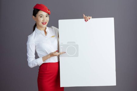 Foto de Feliz moderno asiático femenino vuelo asistente en rojo falda y sombrero uniforme con blanco cartel aislado en gris. - Imagen libre de derechos