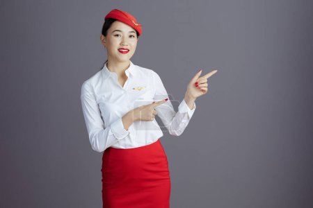 Photo for Happy stylish flight attendant asian woman in red skirt and hat uniform pointing at something isolated on gray background. - Royalty Free Image