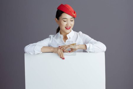 Téléchargez les photos : Souriant élégant air hôtesse asiatique femme en rouge jupe et chapeau uniforme avec blanc panneau d'affichage isolé sur gris. - en image libre de droit