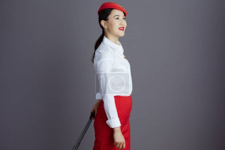 Foto de Sonriente elegante asiática azafata de vuelo en falda roja y sombrero uniforme con bolsa de carro mirando a la distancia aislada sobre fondo gris. - Imagen libre de derechos