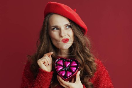 Foto de Feliz San Valentín. hembra pensativa de moda con pelo largo ondulado en suéter rojo y boina y caja de dulces en forma de corazón. - Imagen libre de derechos