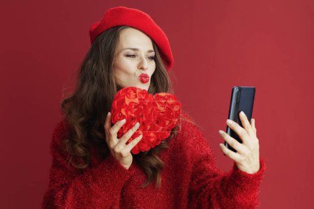 Foto de Feliz San Valentín. mujer de mediana edad de moda en suéter rojo y boina con corazón rojo y teléfono inteligente video chat y envío de un beso. - Imagen libre de derechos