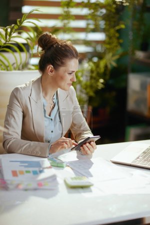 Téléchargez les photos : Femme comptable moderne dans un costume d'affaires léger dans un bureau vert moderne en utilisant smartphone. - en image libre de droit