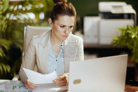 Photo for Pensive modern 40 years old small business owner woman in a light business suit in modern green office with documents and laptop. - Royalty Free Image