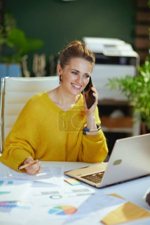 Photo for Happy modern middle aged small business owner woman in yellow sweater with laptop talking on a smartphone and working with documents in the modern green office. - Royalty Free Image