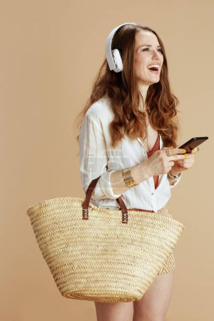 Photo for Beach vacation. smiling elegant middle aged housewife in white blouse and shorts isolated on beige background with straw bag listening to the music with headphones and using smartphone. - Royalty Free Image