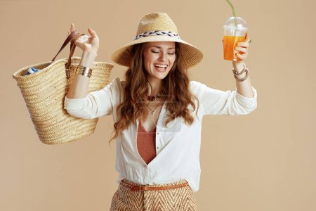 Photo for Beach vacation. happy stylish middle aged housewife in white blouse and shorts isolated on beige background with straw bag, carrot smoothie and summer hat. - Royalty Free Image