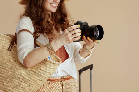 Téléchargez les photos : Vacances à la plage. heureuse femme élégante en chemisier blanc et short isolé sur fond beige avec sac en paille, sac trolley et appareil photo. - en image libre de droit