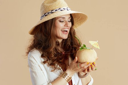 Foto de Vacaciones en playa. elegante mujer de mediana edad sonriente en blusa blanca y pantalones cortos contra fondo beige con cóctel de coco y sombrero de paja. - Imagen libre de derechos