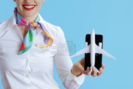 Foto de Primer plano de la azafata moderna feliz sobre fondo azul en uniforme que muestra un pequeño avión y teléfono inteligente. - Imagen libre de derechos