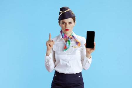 Photo for Modern female air hostess on blue background in uniform showing smartphone blank screen with raised finger drawing attention. - Royalty Free Image