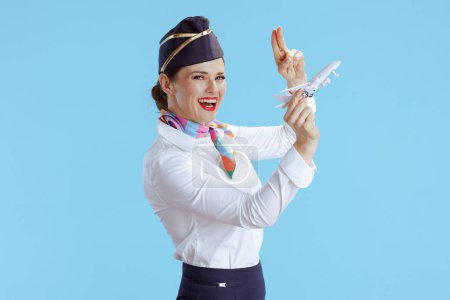 Téléchargez les photos : Heureuse hôtesse de l'air élégante femme sur fond bleu en uniforme avec un petit avion salue. - en image libre de droit