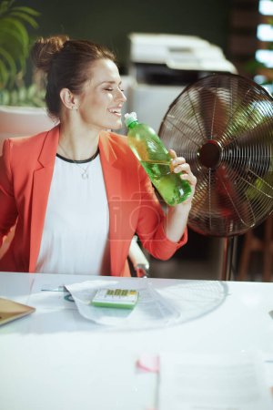 Téléchargez les photos : Un lieu de travail durable. heureuse femme d'affaires moderne dans le bureau vert moderne en veste rouge avec bouteille d'eau et ventilateur électrique. - en image libre de droit