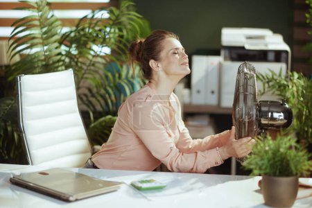Téléchargez les photos : Un lieu de travail durable. heureuse employée moderne dans un bureau vert moderne avec ventilateur électrique. - en image libre de droit