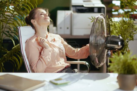 Foto de Lugar de trabajo sostenible. relajado moderno dueño de la pequeña empresa mujer en el trabajo con ventilador eléctrico. - Imagen libre de derechos