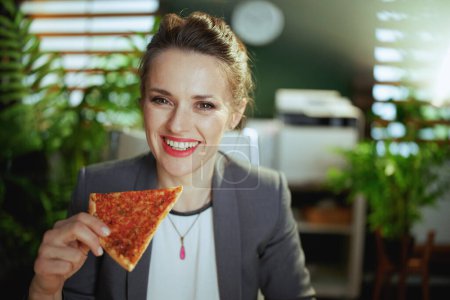 Téléchargez les photos : Un lieu de travail durable. employée moderne souriante dans un costume d'affaires gris dans un bureau vert moderne avec pizza. - en image libre de droit