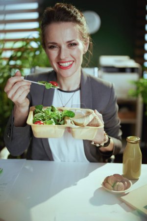 Foto de Lugar de trabajo sostenible. feliz mujer trabajadora moderna en un traje de negocios gris en la oficina verde moderna comer ensalada. - Imagen libre de derechos