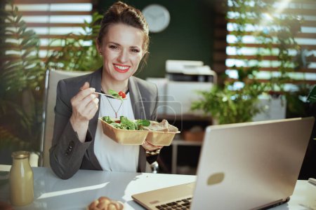 Foto de Lugar de trabajo sostenible. sonriente mujer de negocios moderna en un traje de negocios gris en la oficina verde moderna con portátil comer ensalada. - Imagen libre de derechos