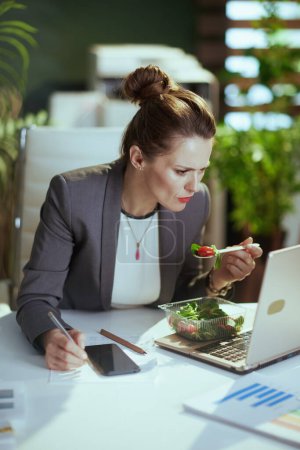 Photo for Sustainable workplace. concerned modern 40 years old business woman in a grey business suit in modern green office with laptop eating salad and using smartphone applications. - Royalty Free Image