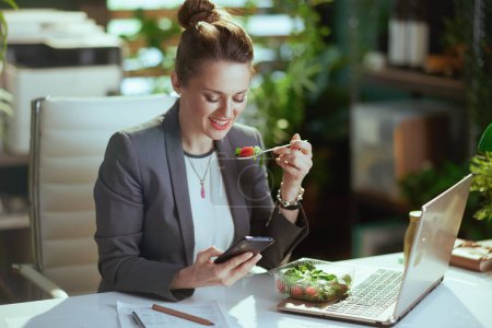Téléchargez les photos : Un lieu de travail durable. heureuse femme d'affaires moderne de 40 ans dans un costume d'affaires gris dans un bureau vert moderne avec ordinateur portable manger de la salade et en utilisant des applications smartphone. - en image libre de droit