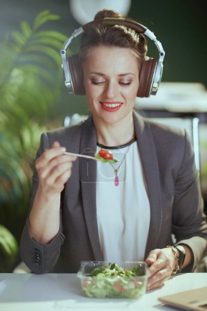 Photo for Sustainable workplace. happy modern accountant woman in a grey business suit in modern green office with headphones eating salad. - Royalty Free Image
