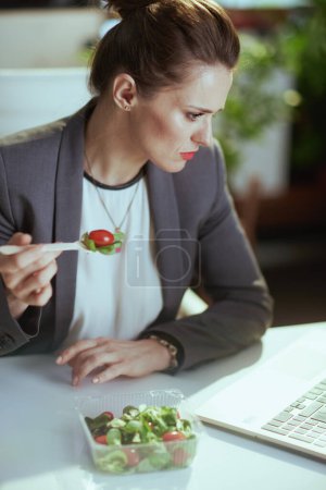 Téléchargez les photos : Un lieu de travail durable. concerné femme comptable moderne de 40 ans dans un costume d'affaires gris dans un bureau vert moderne avec ordinateur portable manger salade. - en image libre de droit