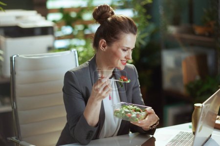 Téléchargez les photos : Un lieu de travail durable. heureuse femme comptable d'âge moyen moderne dans un costume d'affaires gris dans un bureau vert moderne avec ordinateur portable manger de la salade. - en image libre de droit