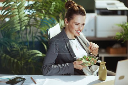 Foto de Lugar de trabajo sostenible. sonriente moderna 40 años de edad dueño de una pequeña empresa mujer en un traje de negocios gris en la oficina verde moderna comer ensalada. - Imagen libre de derechos
