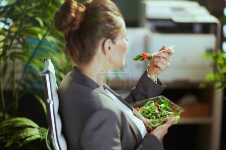 Foto de Lugar de trabajo sostenible. Visto desde atrás mujer dueña de una pequeña empresa de mediana edad en un traje de negocios gris en una oficina verde comiendo ensalada. - Imagen libre de derechos