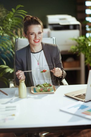 Téléchargez les photos : Un lieu de travail durable. souriant moderne 40 ans femme d'affaires dans un costume d'affaires gris dans un bureau vert moderne avec ordinateur portable manger salade. - en image libre de droit