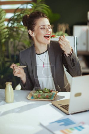 Téléchargez les photos : Un lieu de travail durable. travailleuse moderne souriante dans un costume d'affaires gris dans un bureau vert moderne avec salade mangeant ordinateur portable. - en image libre de droit