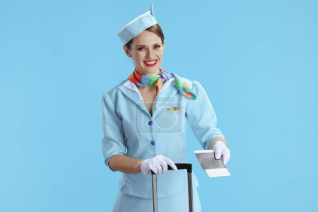 Téléchargez les photos : Hôtesse de l'air féminine élégante souriante sur fond bleu en uniforme bleu avec sac à roulettes, billet et passeport. - en image libre de droit