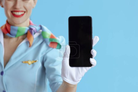Foto de Primer plano de la azafata sonriente del aire femenino sobre fondo azul en uniforme azul mostrando la pantalla en blanco del teléfono inteligente. - Imagen libre de derechos
