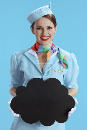 Photo for Happy stylish flight attendant woman isolated on blue background in blue uniform showing blank cloud shape board. - Royalty Free Image