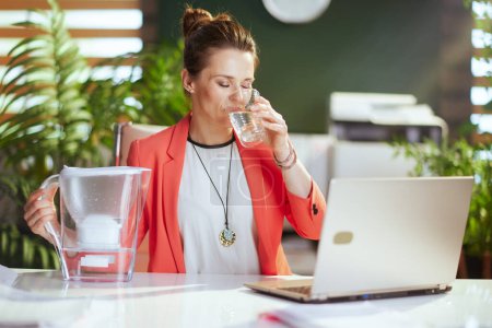 Téléchargez les photos : Un lieu de travail durable. femme d'affaires moderne dans un bureau vert moderne dans une veste rouge avec tasse d'eau, filtre à eau et ordinateur portable. - en image libre de droit