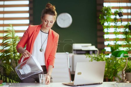 Nachhaltiger Arbeitsplatz. moderne Geschäftsfrau bei der Arbeit in roter Jacke mit Tasse Wasser, Wasserfilter und Laptop.
