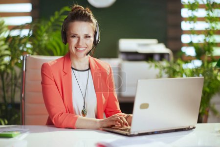 Photo for Sustainable workplace. Portrait of happy modern business woman in modern green office in a red jacket with headset and laptop. - Royalty Free Image