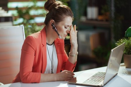 Foto de Lugar de trabajo sostenible. mujer de negocios moderna estresada en el trabajo en una chaqueta roja con auriculares y portátil. - Imagen libre de derechos