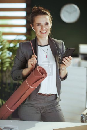 Photo for Sustainable workplace. Portrait of smiling modern small business owner woman in modern green office with wireless headphones, yoga mat and smartphone. - Royalty Free Image