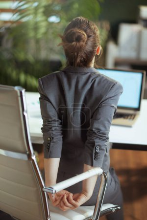 Foto de Lugar de trabajo sostenible. Visto desde atrás mujer de negocios moderna en un traje de negocios gris en la moderna oficina verde estiramiento de la mano. - Imagen libre de derechos