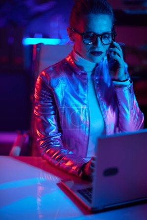 Foto de Concepto futurista neón metaverso. mujer de mediana edad con estilo en gafas con portátil hablando en un teléfono inteligente en la oficina moderna. - Imagen libre de derechos